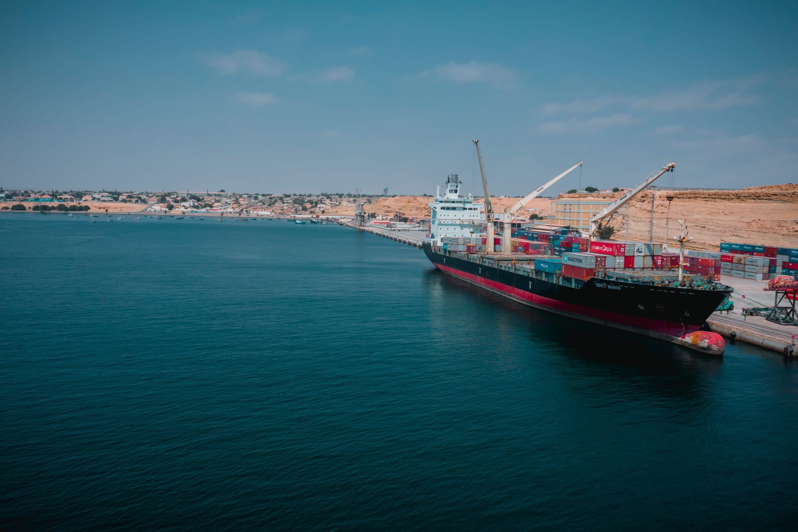 A container ship alongside a quay at a coastal terminal, cranes working the deck, seen across calm water.