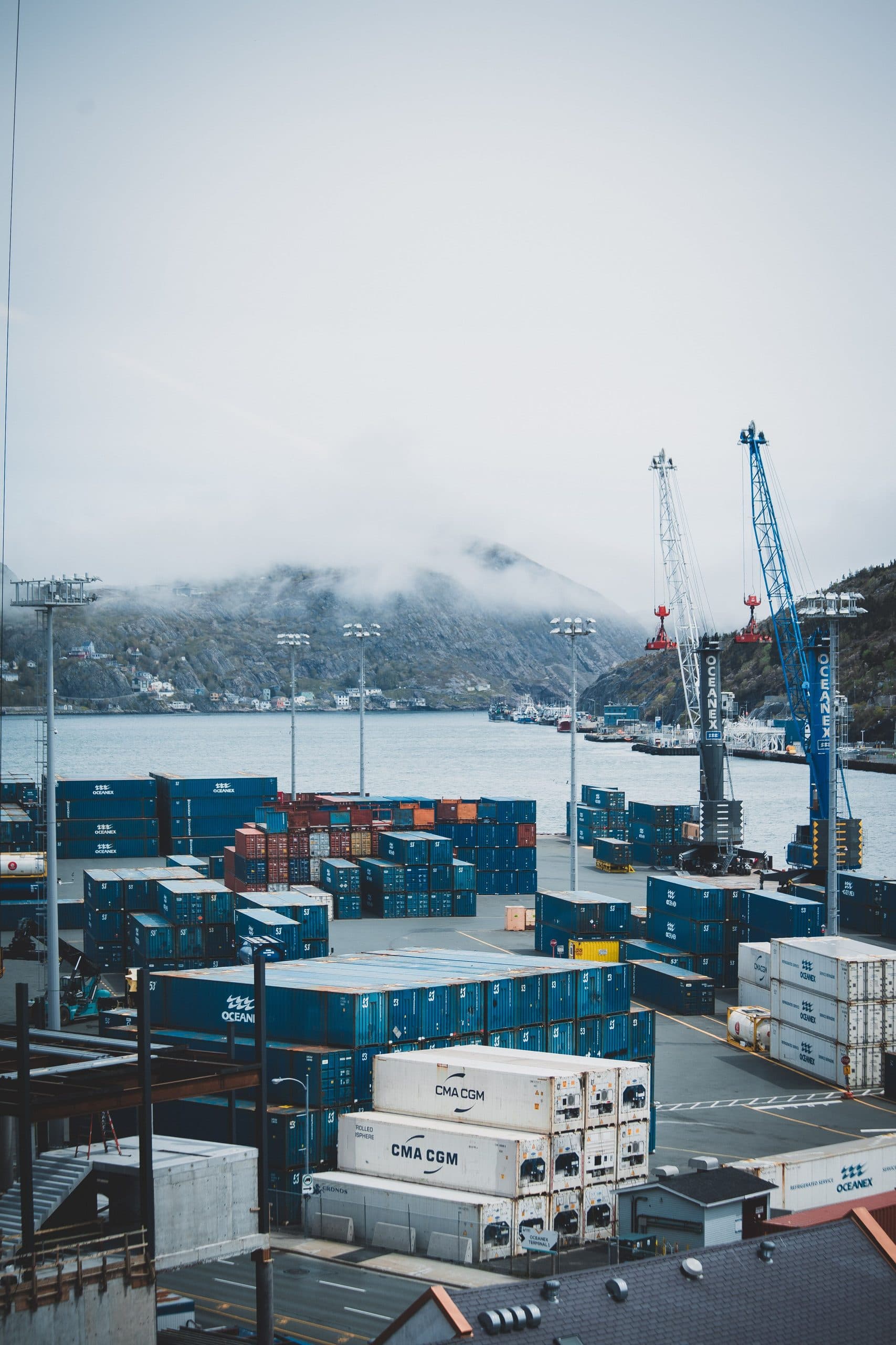 Stacked shipping containers in a terminal yard, with gantry cranes behind them and low cloud on the hills beyond.