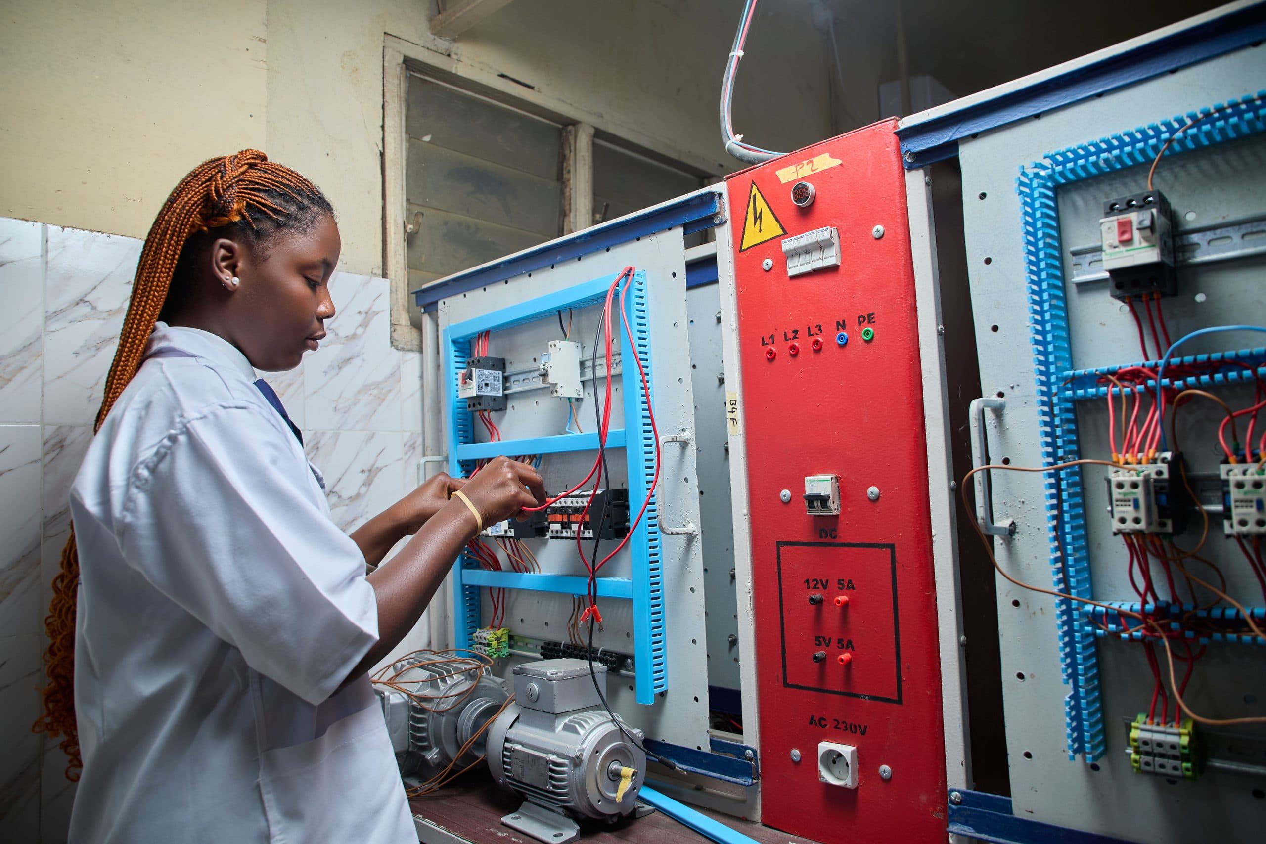 A technician in a white coat working into an open electrical control panel, holding a test probe against the wiring.