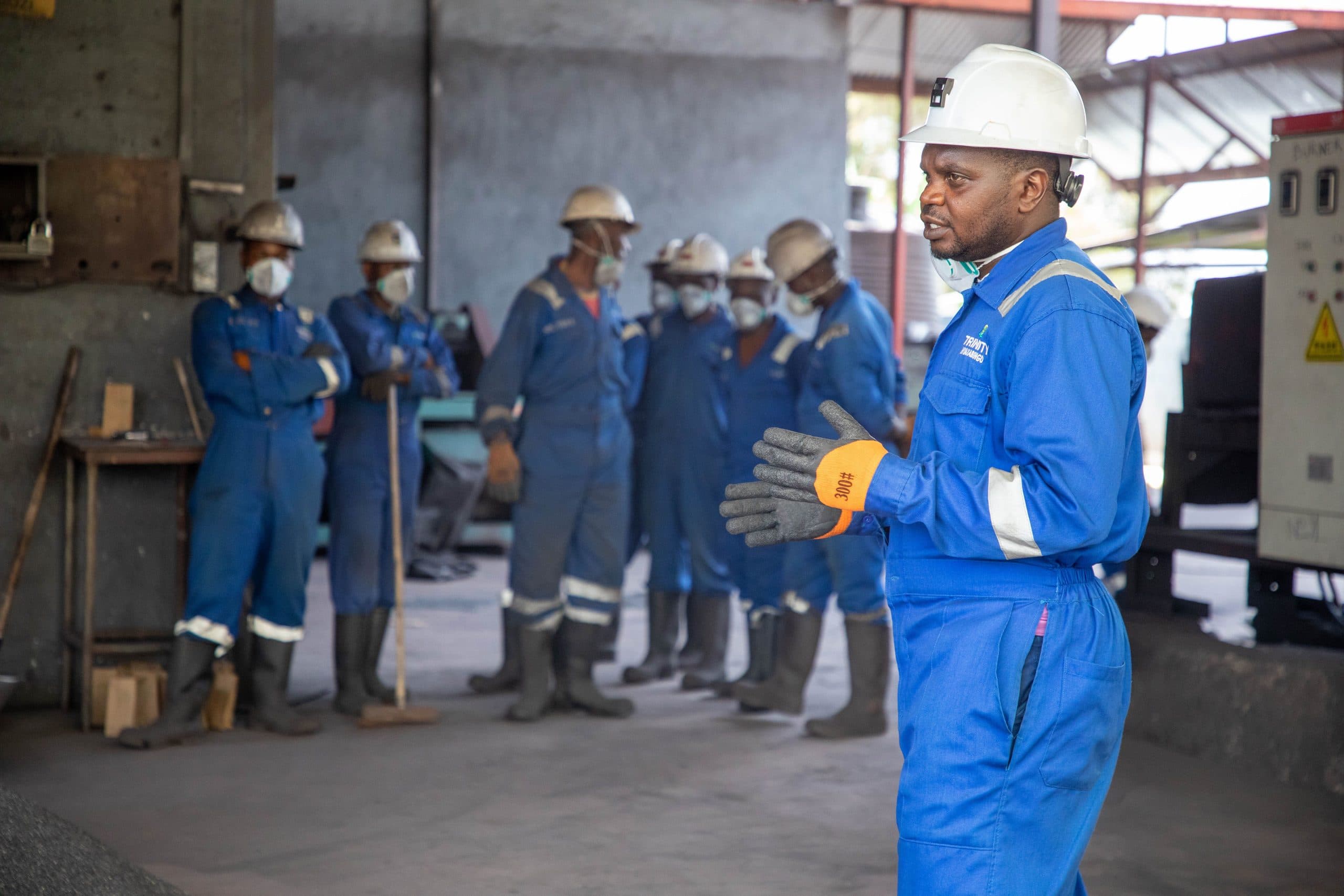 An instructor in blue coveralls briefing a line of workers in hard hats and high-visibility gear inside an industrial building.