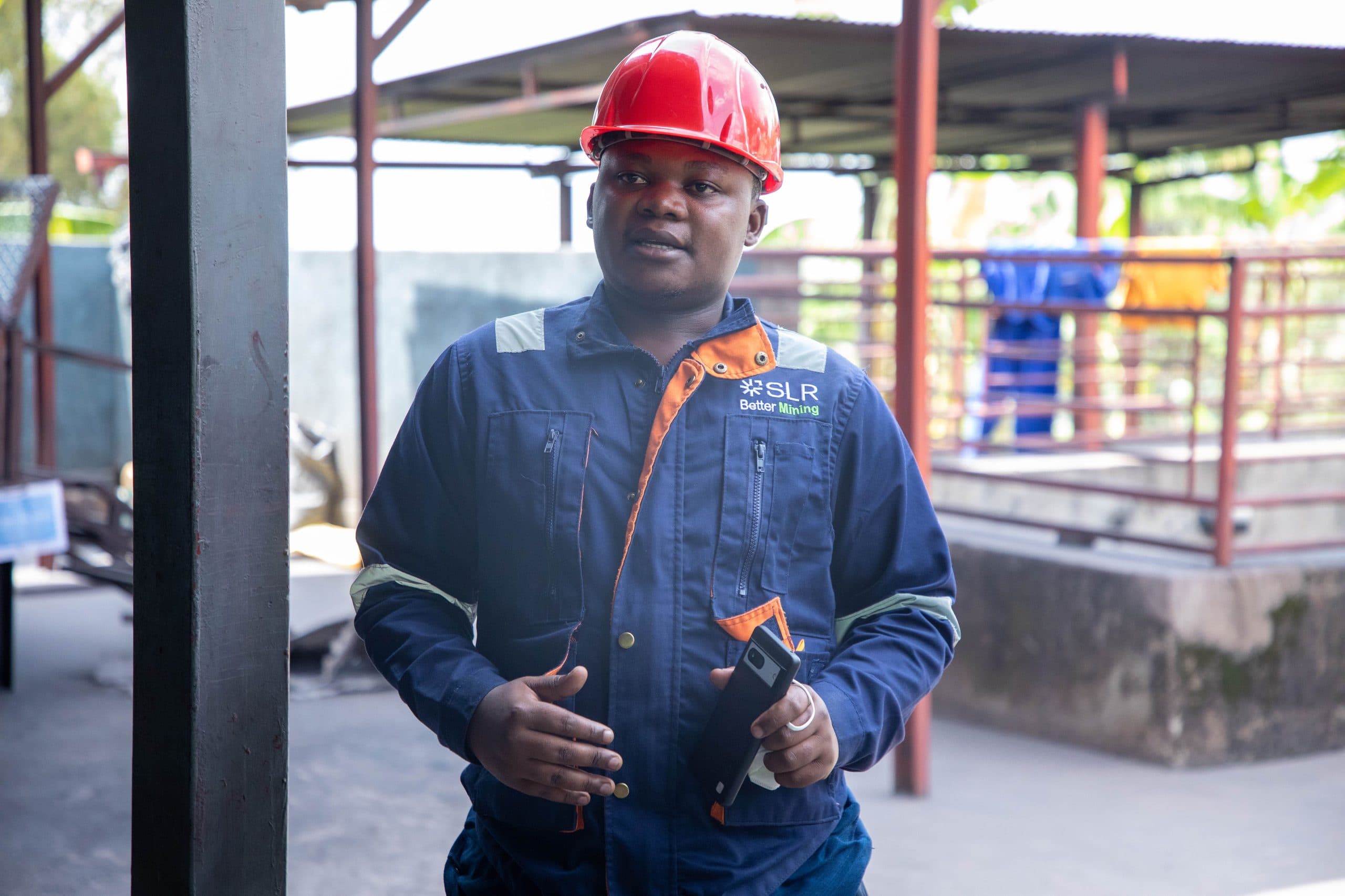An engineer in coveralls and a hard hat standing on an industrial site, holding a handheld device.