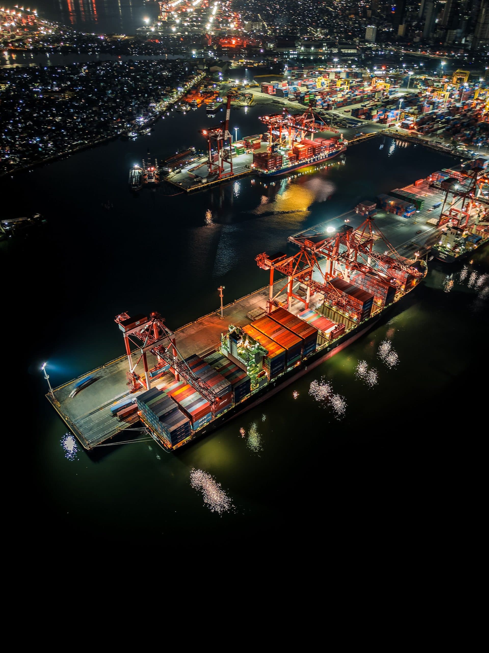 A container terminal seen from the air at night, quays and stacked containers lit by floodlights against dark water.