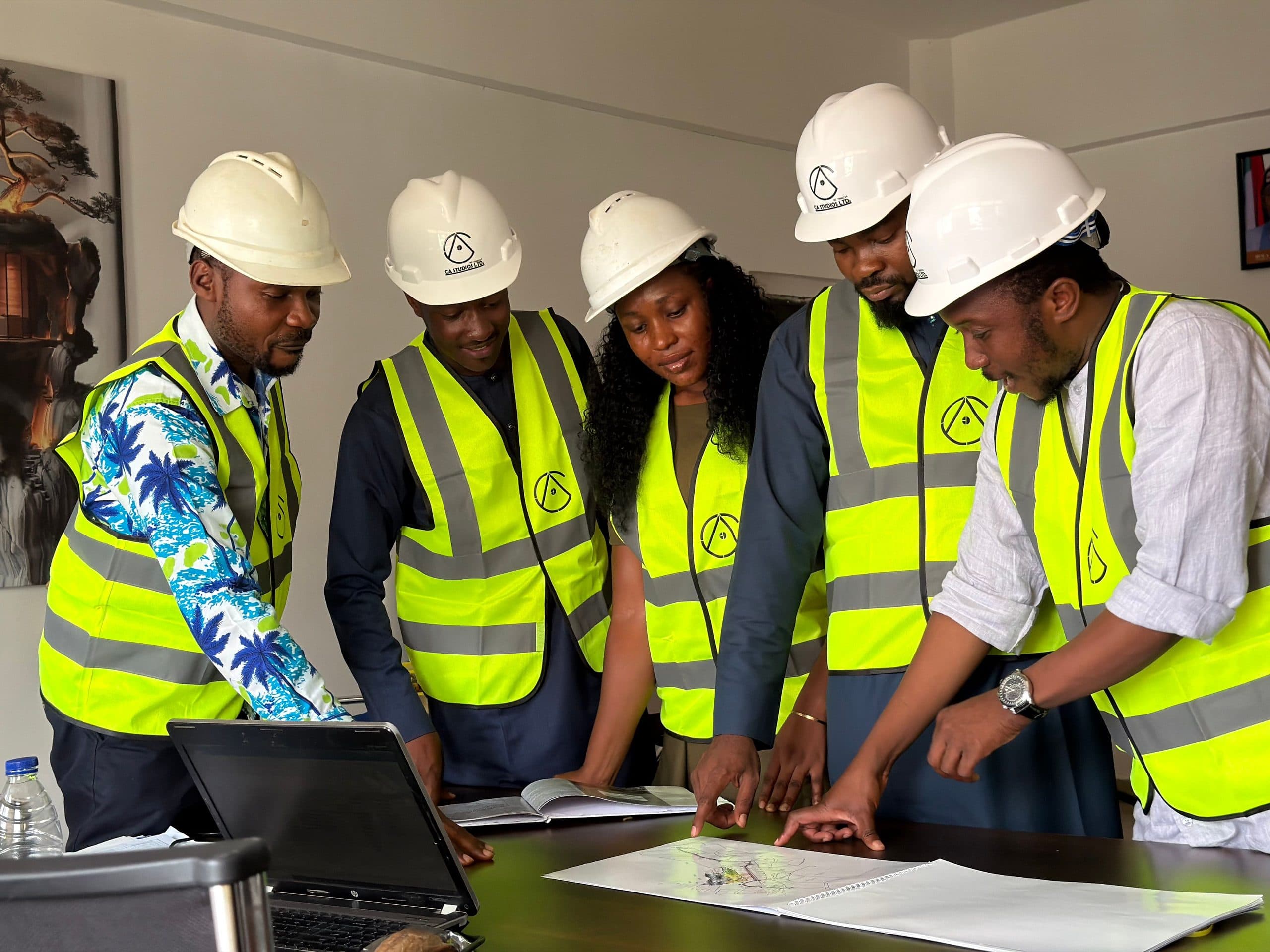 A team of engineers in hard hats and high-visibility vests gathered around a drawing and a laptop, discussing it.