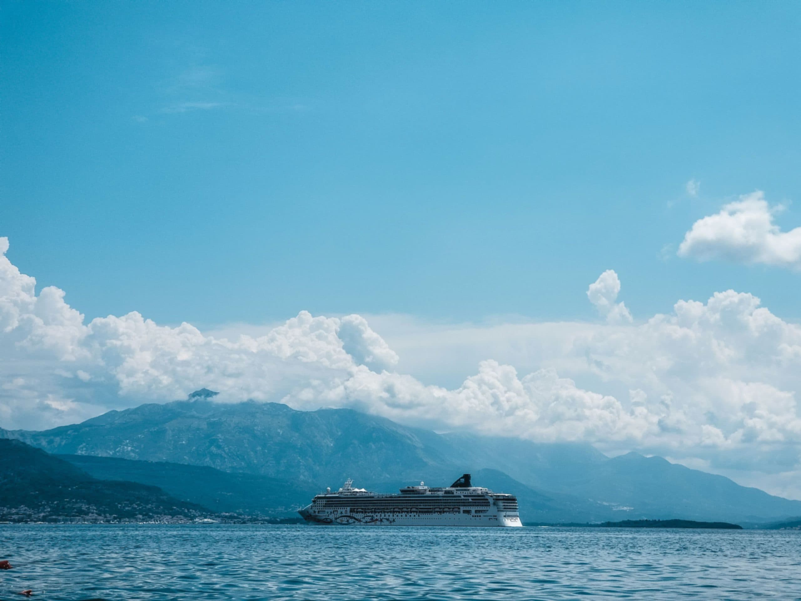 A vessel moving along a coastline with mountains rising behind it under a bright sky.