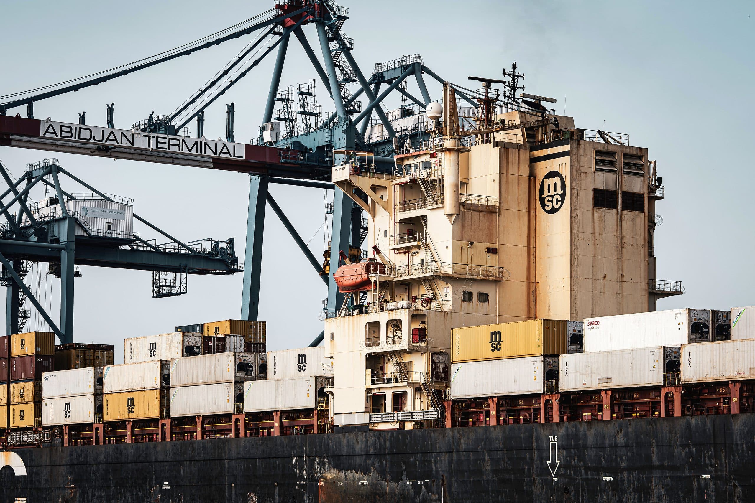 Container cranes and stacked containers at a West African terminal, with a cargo vessel alongside the quay.
