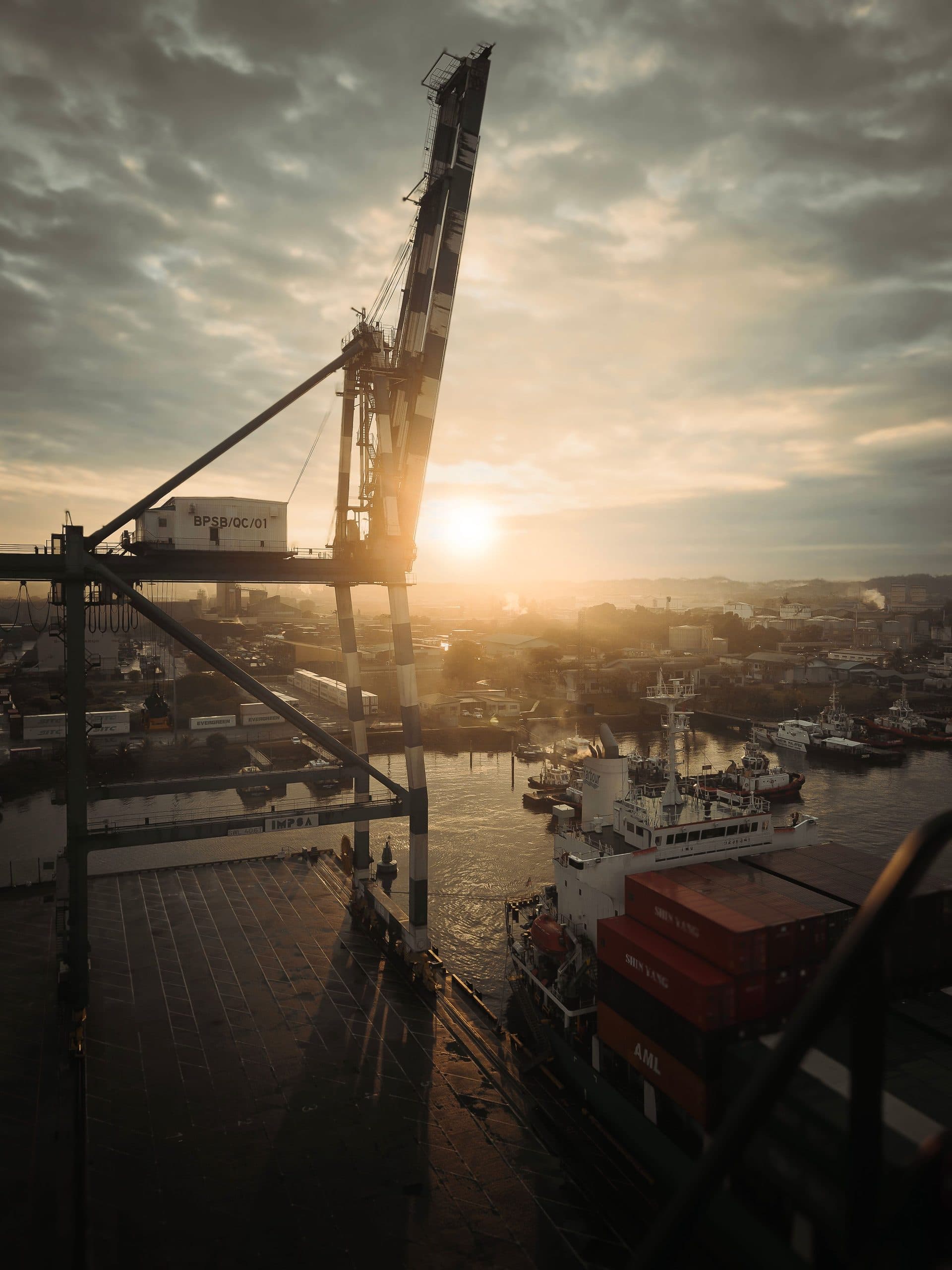 A quayside gantry crane silhouetted against a low sun, with shipping containers stacked beneath it.