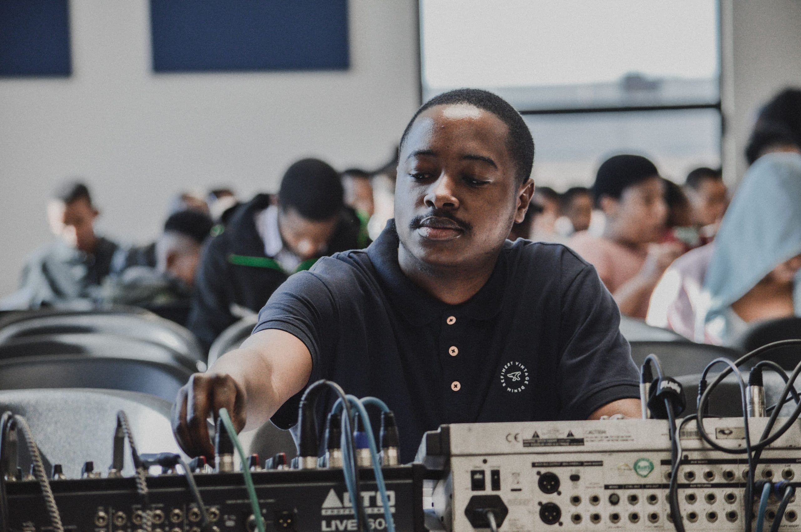 A man working at an audio control desk, with rows of seated people filling the hall behind him.